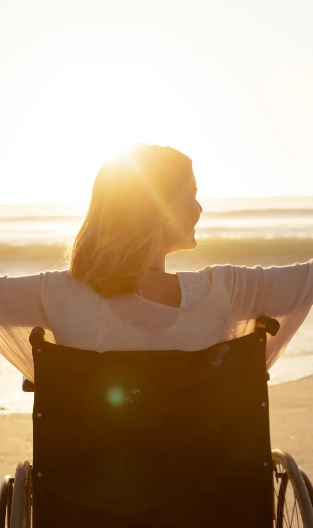A photo of a woman sitting in a wheelchair with her arms extended to her sides, basking in the sunshine of a sunset on the beach.