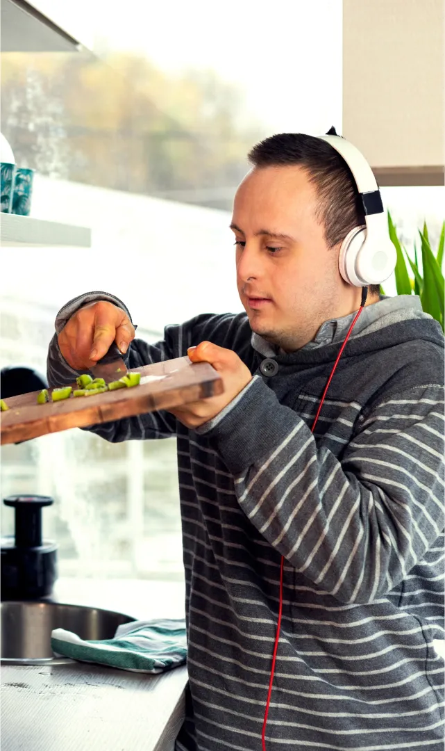 A photo of a man with down syndrome wearing headphones and cooking in his kitchen, scraping vegetables into a pot.