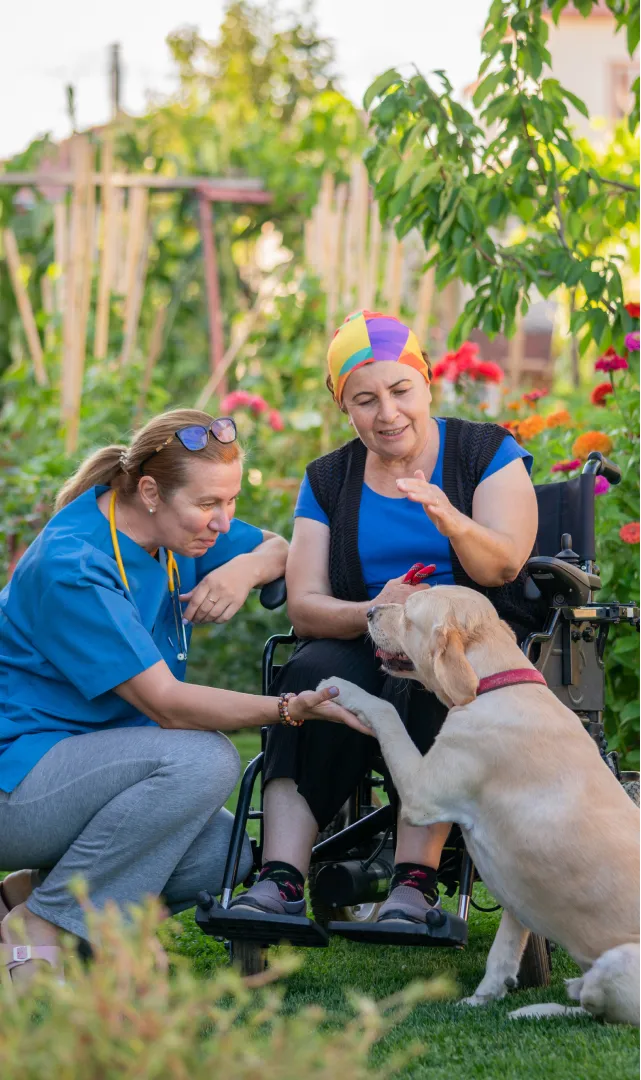 A photo of a woman in a wheelchair with her carer and a golden retriever, sitting in her backyard with lush greenery around.